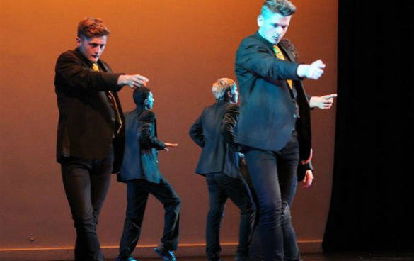 Four teenage boys in black suits stepping forwards pointing their index finger.