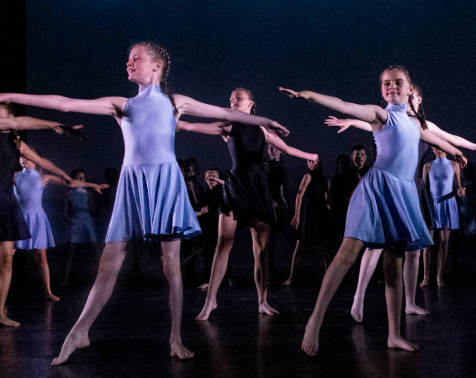 A group of young, female ballet dancers in blue leotards dancing on stage.
